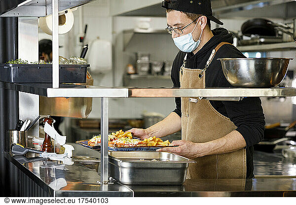 Chef with protective face mask holding food plates at restaurant kitchen