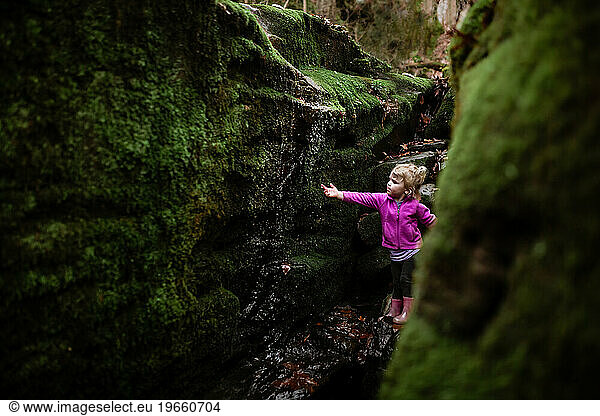 Child holding hand under waterfall surrounded in moss on hike