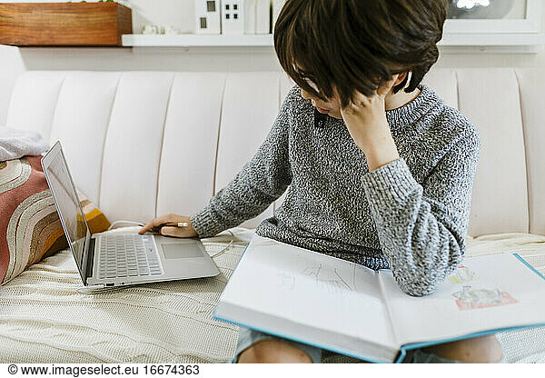 Child sitting on sofa looking at laptop white sketching on a book