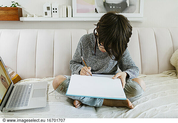 Child sitting with a sketch book on sofa learning remotely how to draw