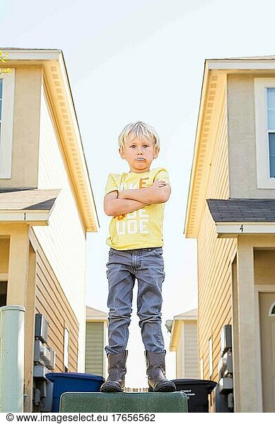 Child standing on a box between townhouses with arms folded