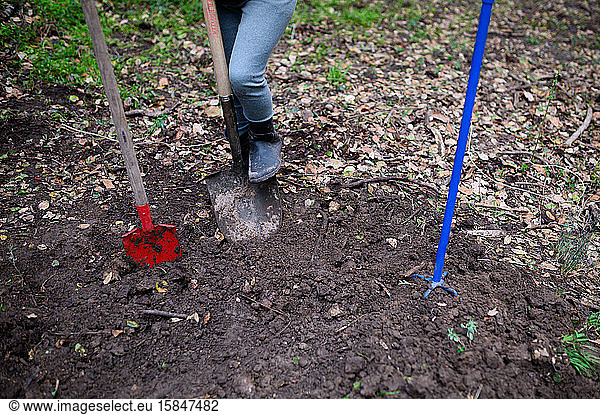 Child wearing boots shoveling soil with other garden tools in yard