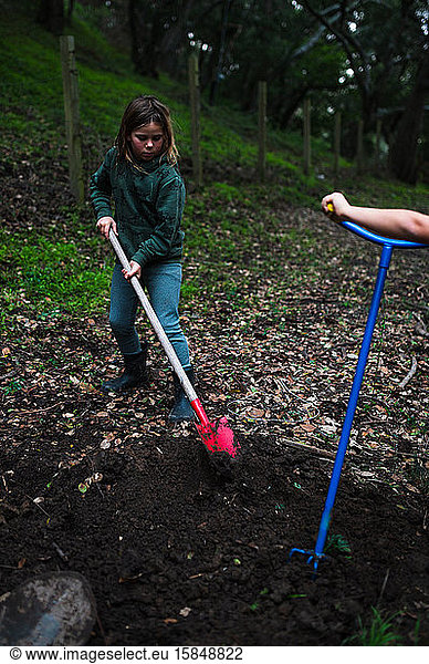 Child with red shovel scooping soil in front yard with brother