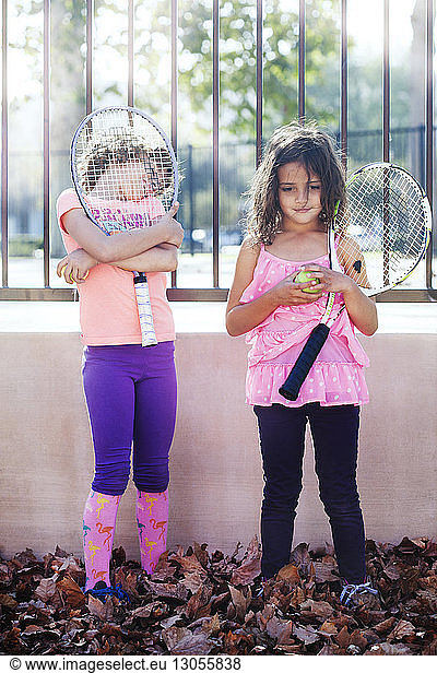 Children holding tennis racket and ball while standing against fence