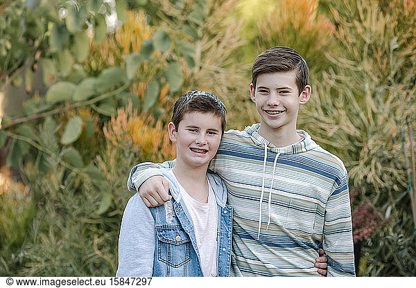 Clean-cut smiling brothers with braces hugging in front of lush plants