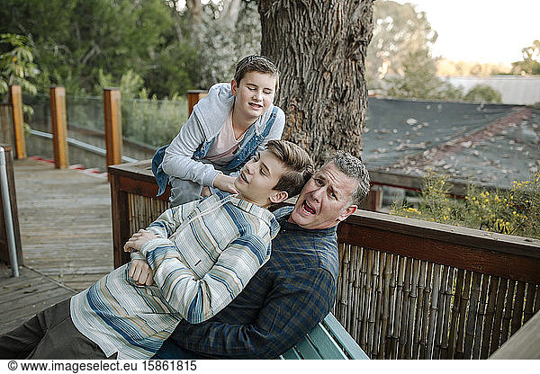 Clean-cut teen sits on dad on bench outdoors with brother watching