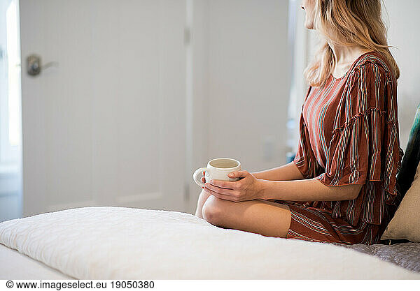Close frame of woman sitting on side of bed holding coffee mug
