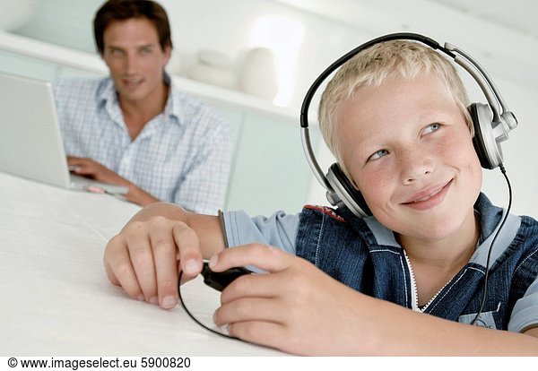 Close_up of a boy wearing headphones and listening to music with his father using a laptop in the background