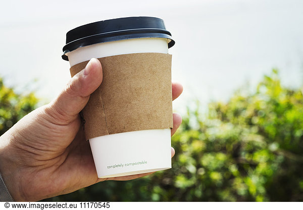 Close up of a human hand holding a disposable paper cup with a cardboard sleeve and plastic lid.