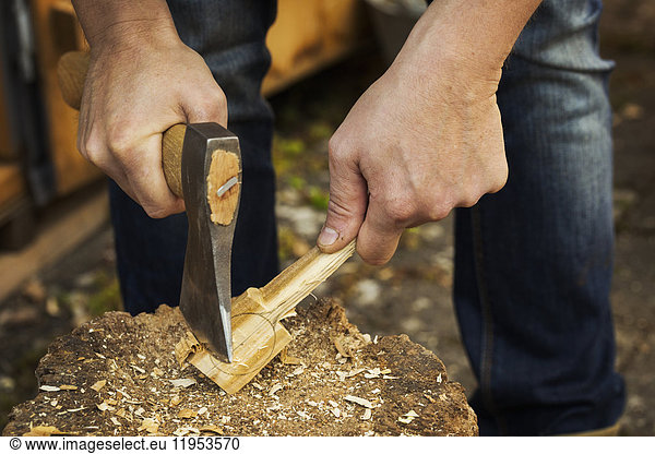 Close up of a man's hand holding an axe  cutting and shaping a small piece of wood on a splitting block covered in wood shavings.