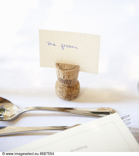 Close up of a place setting at the top table at a wedding banquet. An upside down cork with name tag for the groom. Silver fork and spoon.