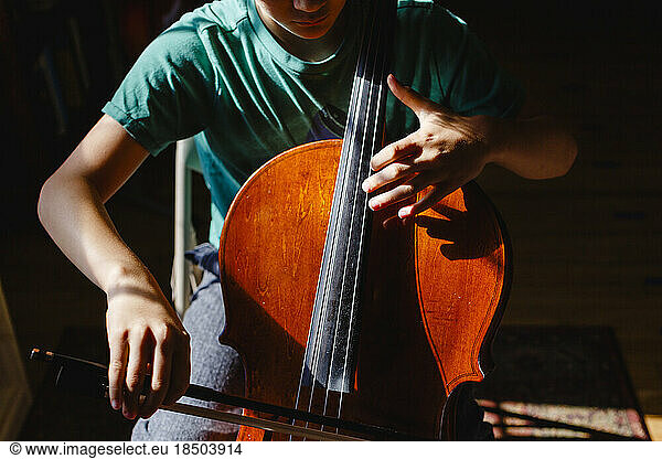 Close-up of boy sitting in bright patch of light practicing cello