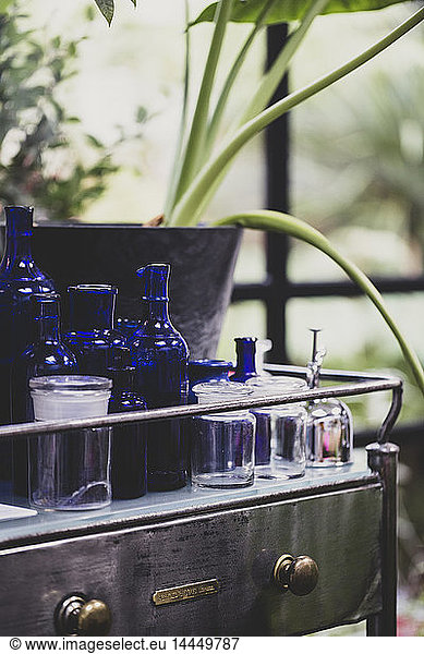Close up of clear and blue glass jars and bottles on vintage metal table with drawer.