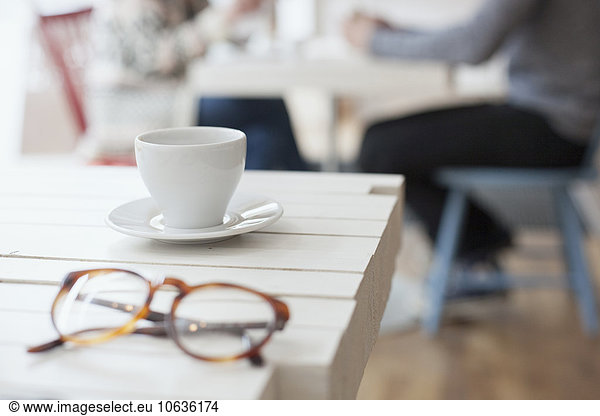Close-up of coffee cup and eyeglasses on cafe table with people in background