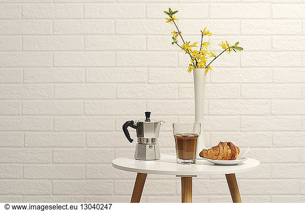 Close-up of croissant with coffee and espresso maker by flower vase on table by wall