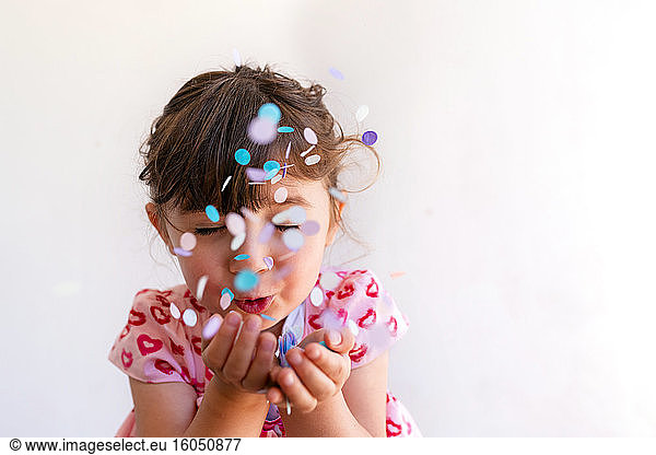 Close-up of cute girl blowing confetti from hands against white background