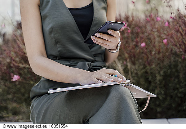 Close-up of female professional with note pad using smart phone while sitting against plants
