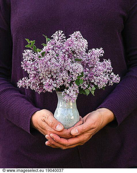 Close up of hands holding a small vase of purple lilac flowers.
