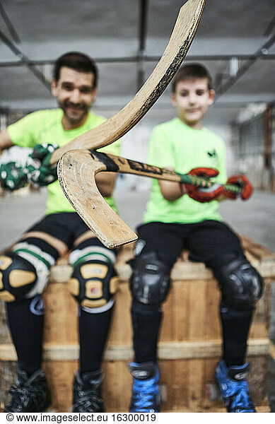 Close-up of hockey sticks held by father and son sitting on wooden box at court