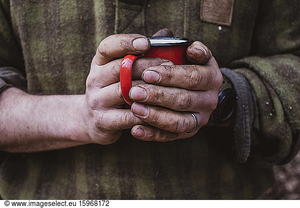 Close up of man with dirty hands holding red enamel mug.