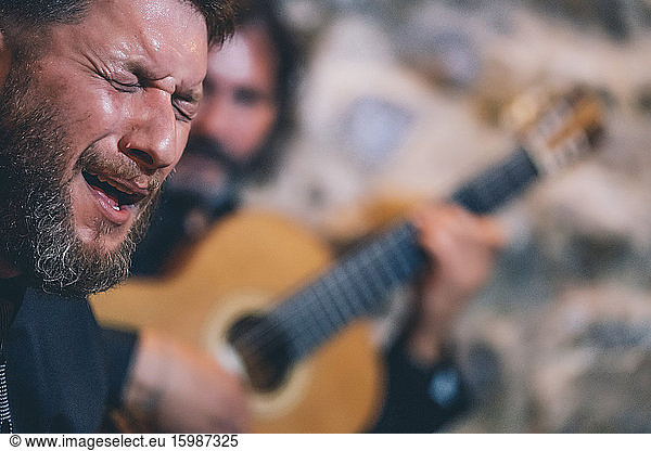 Close-up of man with eyes closed singing while guitarist playing guitar in background