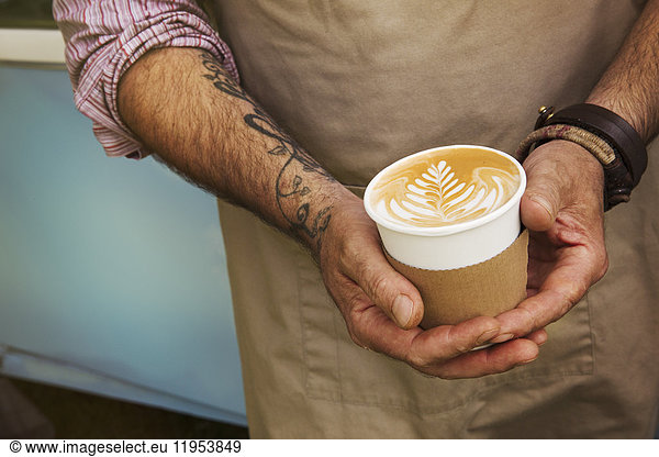 Close up of man with tattoo on his arm holding paper cup with cafe latte.