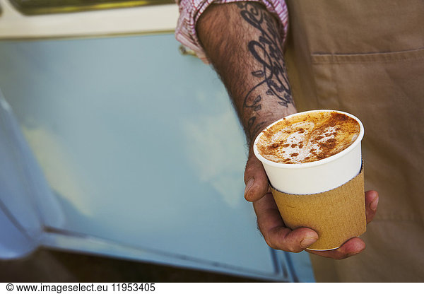 Close up of man with tattoo on his arm holding paper cup with cappuccino.