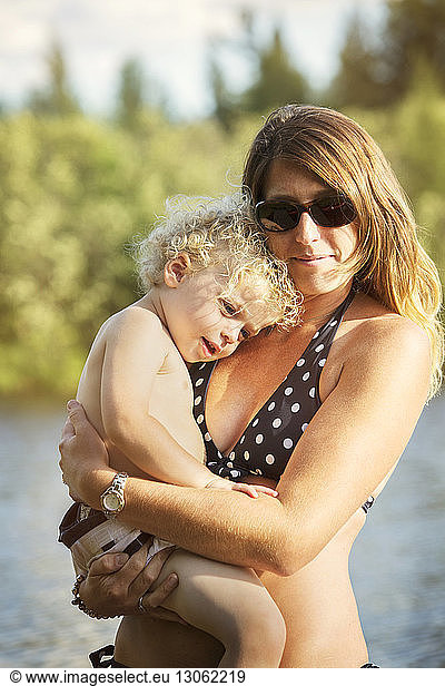 Close-up of mother wearing swimwear carrying smiling son against lake