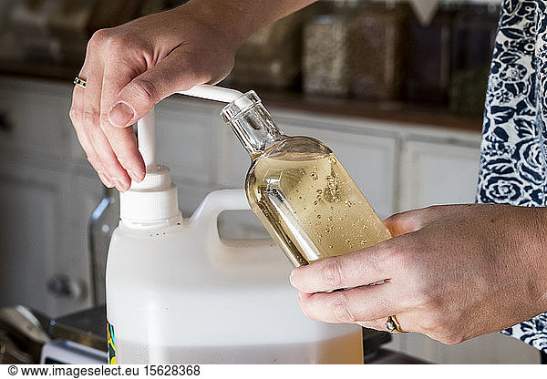 Close up of person standing in a kitchen  decanting liquid from plastic container into glass bottle.