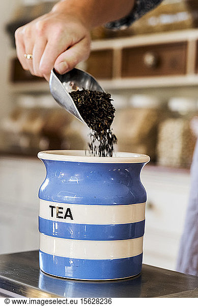 Close up of person standing in a kitchen  placing loose tea into striped blue ceramic jar.