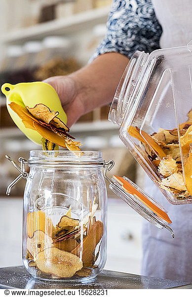 Close up of person standing in a kitchen  placing slices of dried fruit into glass jar.