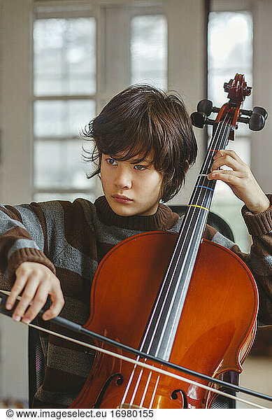 Close-up of preteen boy with serious expression playing cello at home