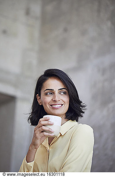 Close-up of smiling female entrepreneur holding coffee cup against wall in office