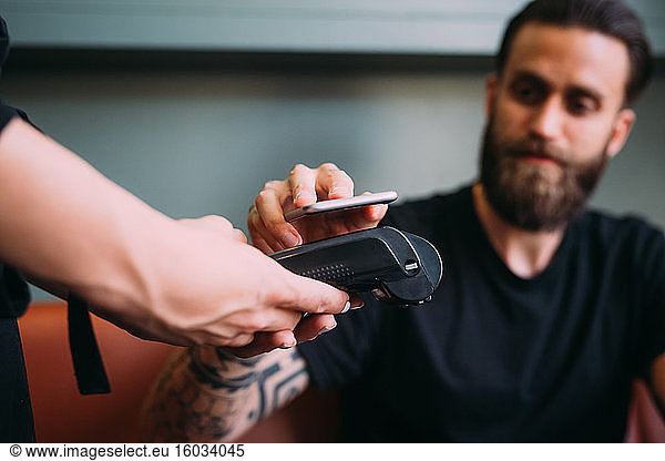 Close up of tattooed bearded man sitting in a bar  using mobile phone to pay.
