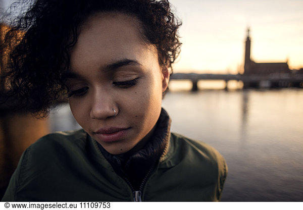 Close-up of teenage girl with curly short hair looking down while standing against canal in city