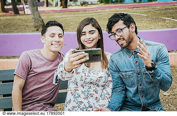 Close up of three friends taking a selfie sitting in the park  Three smiling friends sitting in the park taking a selfie. Front view of three happy friends taking a selfie while sitting on a bench