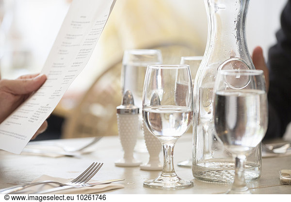 Close up of wine and water glasses and place settings at a table in a restaurant. A person's hand holding the menu.