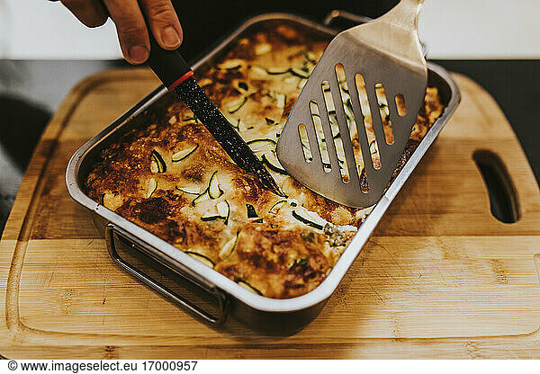 Close-up of woman hand cutting cooked lasagna with zucchini in tray at home