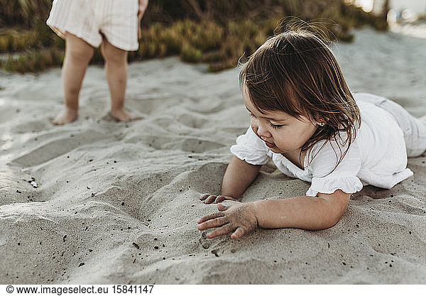 Close up of 2 year old playing in sand at beach while sister looks on