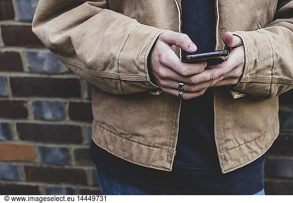 Close up of young man wearing casual jacket standing against a brick wall  texting on his mobile phone.