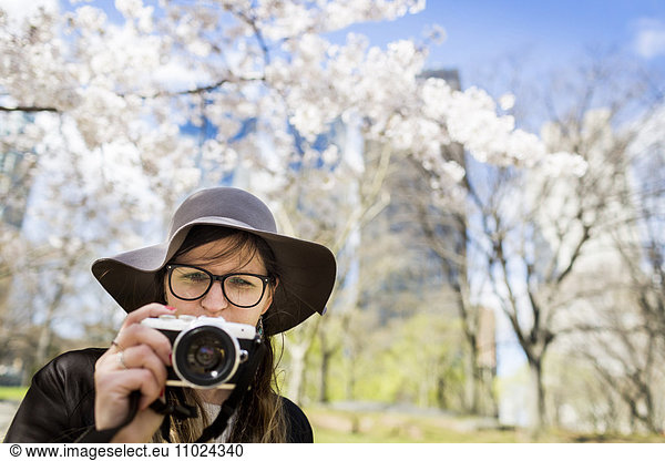 Close-up of young woman with camera standing against trees at Central Park