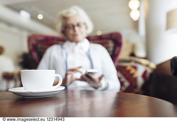 Coffee cup on table and senior woman using mobile phone during coffee break in cafe