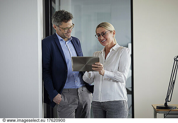 Colleague explaining over tablet PC to businessman leaning on wall with hand in pocket at office