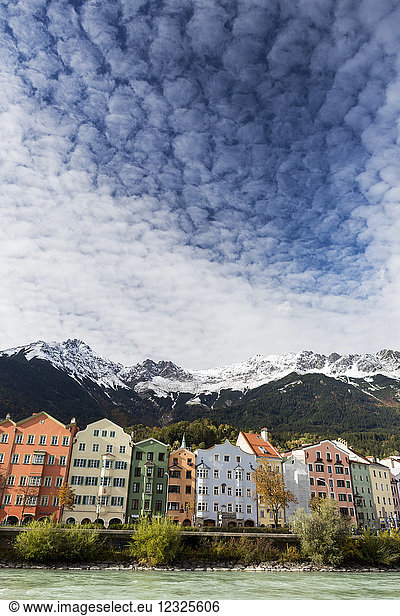 Colourful buildings along river bank with snow-covered mountain peaks  dramatic clouds and blue sky overhead; Innsbruck  Tyrol  Austria