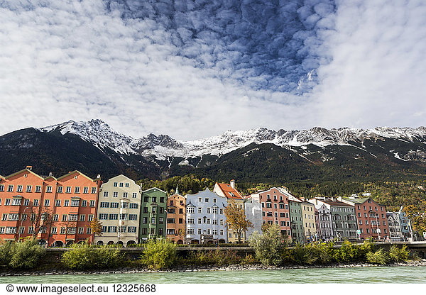 Colourful buildings along river bank with snow-covered mountain peaks  dramatic clouds and blue sky overhead; Innsbruck  Tyrol  Austria