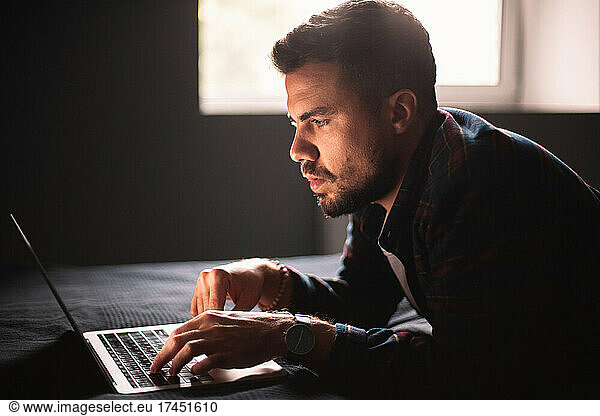 Concentrated man using laptop computer lying on bed at home