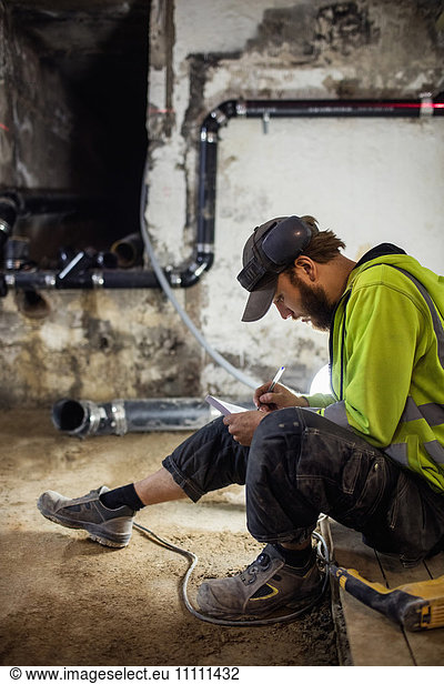 Concentrated manual worker writing notes while drilling floor in basement