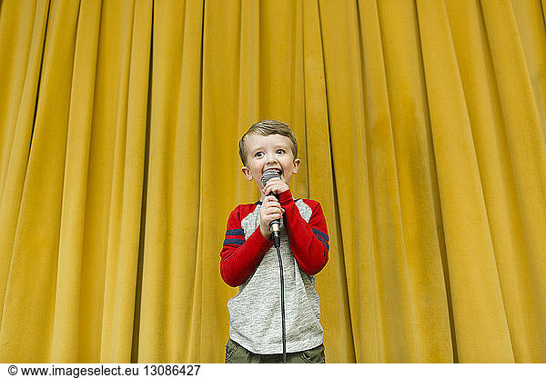 Confident boy singing on microphone at stage against yellow curtains