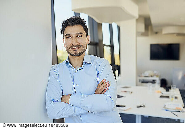 Confident businessman with arms crossed leaning on wall at workplace