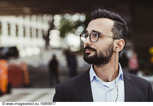 Confident businessman with in-ear headphones looking away while standing outdoors
